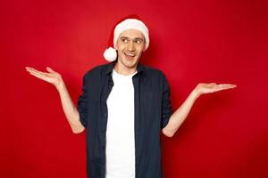 Horizontal christmas shot of attractive man with puzzled expression spreads hands with confused look, he smiles absurdly wears casual clothes feels hesitant isolated over red background. Doubt concept photo