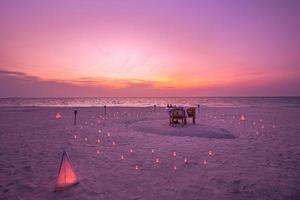 Beautiful table set up for a romantic meal on the beach with lanterns and chairs and flowers with candles and sky and sea in the background. Sunset beach dinner