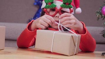menina com chapéu de Papai Noel, abrindo a caixa de presente, sentado na sala de estar com a árvore de Natal em casa. feliz Ano Novo e feliz Natal. video