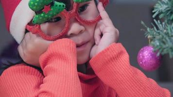 petite fille dans un bonnet de noel assise sur le sol et faisant des voeux près de l'arbre de noël à la maison. Bonne année et joyeux Noël. video