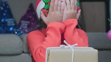 menina com chapéu de Papai Noel, abrindo a caixa de presente, sentado na sala de estar com a árvore de Natal em casa. feliz Ano Novo e feliz Natal. video