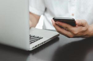 Mano de hombre usando un teléfono inteligente y una computadora portátil en la mesa en casa, buscando información navegando por Internet en la web, trabajando desde casa concepto de compras en línea de negocios foto