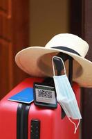 New normal concept. Close up view of a smartphone with an immune digital health passport, a red suitcase, protective mask, straw hat, passport and sunglasses. Traveling by plane during a pandemic. photo