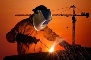 Welding worker welding steel structure with construction crane and building at construction site on sunset for construction industrial work concept. photo