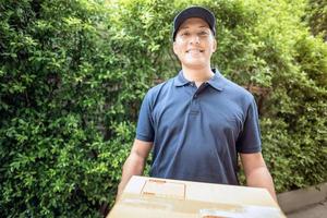 apuesto joven asiático repartidor sonriendo mientras sostiene una caja de cartón a su cliente. foto