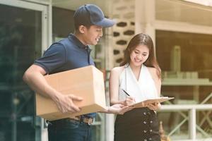 Handsome young asian delivery man smiling and holding a cardboard box while waiting beautiful young asian woman putting signature in clipboard for confirm receive. photo