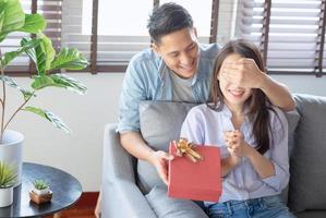 Hombre guapo le está dando a su novia una caja de regalo roja para cumpleaños sorprendente en la sala de estar de casa foto