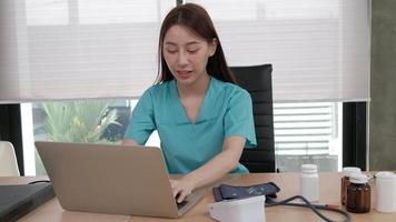 Young beautiful Asian female doctor with stethoscope and uniform working and video chatting with a smile about healthy with laptop computer on a desk, medicine nearby at hospital clinic office.