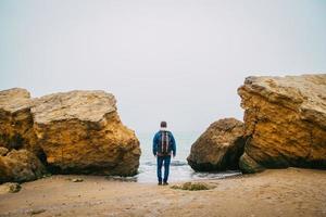 Traveler man with backpack standing on sandy beach in middle of rocks against sea background photo