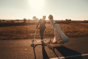 Sonriente pareja de novios montando en scooters a lo largo de la carretera fuera de la ciudad al atardecer. lugar para texto o publicidad foto