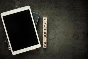 Blank screen tablet with notebook and word e-learning on wood cube block on black table background photo