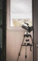Un gran telescopio gris de pie en el balcón apuntaba hacia el cielo nublado a través de la ventana. sin observador del cielo foto