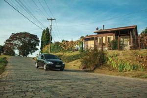 Bento Goncalves, Brazil - July 13, 2019. Car passing by a stone paved road next to a charming wood house near Bento Goncalves. A friendly country town famous for its wine production.