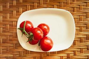 A cluster of red cherry tomatoes on a white flat rectangular plate stand on a wooden wicker surface. View from above. Life style composition.