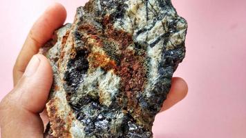 andesita ígnea alterada en zona de alteración hidrotermal, con vetas de cuarzo, clorita y minerales de pirita negra brillante, sobre fondo rosa. indonesia, exploración geológica.