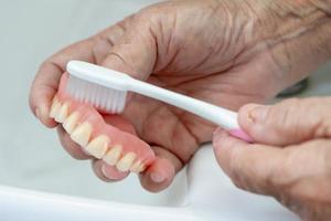 Paciente asiático mayor o anciana usa cepillo de dientes para limpiar la dentadura parcial de los dientes de reemplazo. foto