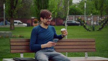 Handsome young man browsing smartphone social network app smiles texting a message while drink coffee. video
