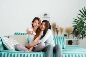LGBT lesbian women couple moments happiness. Lesbian women couple together indoors concept. Friends young smiling women at home sitting on the couch and watching tv, She is holding a remote control. photo