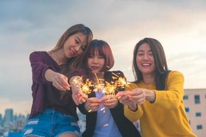 Outdoor shot of young people at rooftop party. Happy group of asia girl friends enjoy and play sparkler at roof top party at evening sunset. Holiday celebration festive party. Teenage lifestyle party. photo
