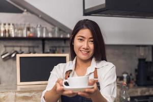 Cerca de una joven barista asiática sosteniendo una taza de café que sirve a su cliente con una sonrisa rodeada de fondo de barra de bar. joven barista y su pequeña tienda. concepto de comida y bebida foto