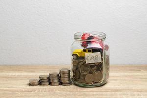 Moneda y coches en un frasco de vidrio para ahorrar dinero en la mesa con fondo blanco. foto