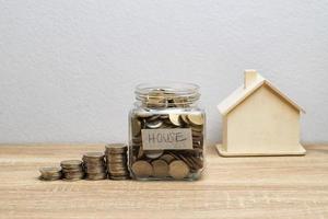 Moneda en un frasco de vidrio con pila de dinero y casa de madera blanca sobre la mesa marrón foto