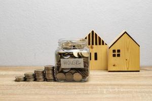 Moneda en un frasco de vidrio con pila de dinero y casas de madera marrón sobre la mesa foto