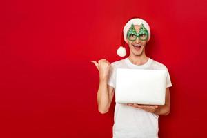 excited man with laptop in hand points with thumb vacant empty space for advertising. dressed Santa Christmas hat. isolated on red background. concept - people, technology, discounts, online shopping photo