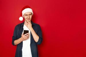 young man with phone in hand smiles slyly, thinks about new idea, congratulations. Santa Claus Christmas hat. isolated on red studio background space for text. concept - people, technology, holiday photo
