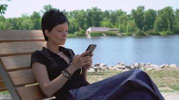 garota conversando na célula sentada perto do rio. video