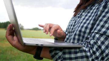 gros plan sur les mains femme asiatique aux cheveux noirs debout et travaillant sur un ordinateur portable dans une ferme, marchant et vérifiant autour de la ferme. grands arbres et fond d'herbe verte. être dans une belle ferme verte video