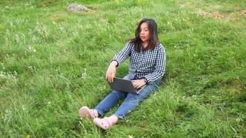 mujer asiática trabajando en la computadora portátil y tomando un descanso en una granja. trabajando entre la hermosa vista de la naturaleza, acostado y poniendo la mano sobre la hierba verde. trabajando en un concepto de granja video