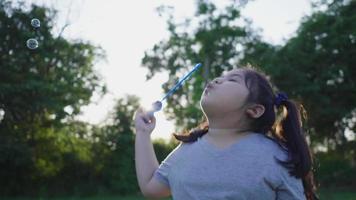 menina asiática soprando bolha no parque, jogando bolha no fim de semana, tomando um pouco de ar fresco de grandes árvores no parque, passando o tempo nas férias no parque. fundo de luz solar video