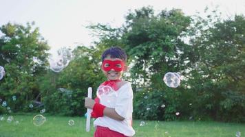 framifrån av glad asiatisk pojke som bär hjältedräkt som spelar bubblor i parken, tillbringar tid på semestern i parken. vacker solljus bakgrund video