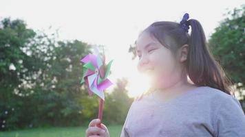 niña asiática feliz del niño que se divierte con el juguete del molino de viento en el parque, alegre y refrescante. concepto de relajación de ocio en vacaciones. hermoso fondo de la luz del sol video