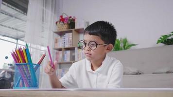 Niño feliz con gafas asiáticas pintando en un libro para colorear mientras está sentado en el piso en la sala de estar. feliz niño tailandés dibujando en papel en vacaciones en casa. concepto de relajación y afición. video