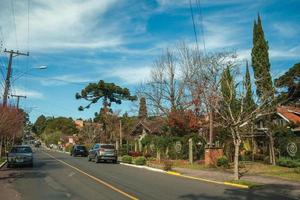 Gramado, Brazil - July 21, 2019. Gardened houses and trees on empty paved street with parked cars in a sunny day at Gramado. A cute european-influenced town highly sought after by tourists. photo