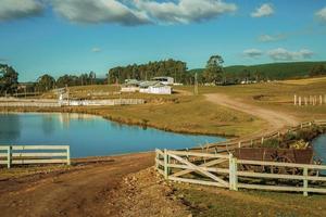 Cambara do Sul, Brazil - July 18, 2019. Pretty farm with fences, livestock sheds and blue lake on landscape of rural lowlands called Pampas at Cambara do Sul.