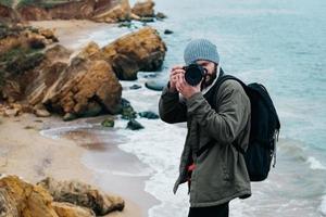 Hombre fotógrafo viajero con mochila tomando fotografías sobre el mar y las rocas de fondo foto