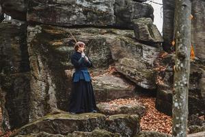 Hombre en kimono negro practicando artes marciales con una espada sobre las rocas y el fondo del bosque foto