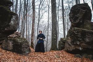 Hombre en kimono negro practicando artes marciales con una espada sobre las rocas y el fondo del bosque foto