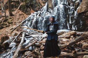 Hombre practicando kendo con espada de bambú en cascada, rocas y fondo de bosque foto