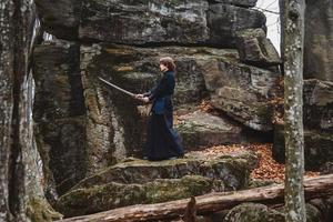 Hombre en kimono negro practicando artes marciales con una espada sobre las rocas y el fondo del bosque foto