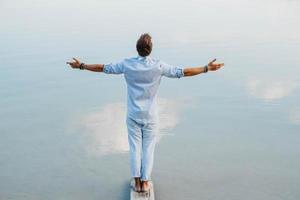 Man handsome stands on wooden bridge and looks at water reflex of blue sky flinging his hands to side photo