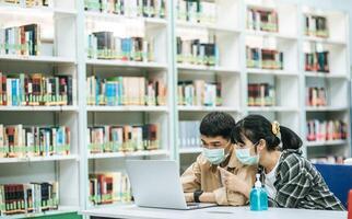 hombres y mujeres usan máscaras y usan una computadora portátil para buscar libros en la biblioteca. foto