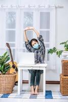 A woman wearing a mask uses a laptop at work and stretches her arms to relax. photo