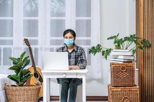 una mujer que lleva una máscara de higiene está sentada en el escritorio con una computadora portátil.