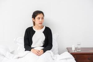 An uncomfortable woman sits on the bed and has medicine on the table. photo