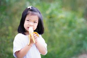 Asian kid 4 year old eating banana during day. Sweet fruit. Empty space.