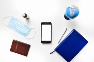 Top view flat lay mobile phone green screen copypaste with blue notebook, passport, sanitizer, blue face mask, global stand on white table isolated background. photo